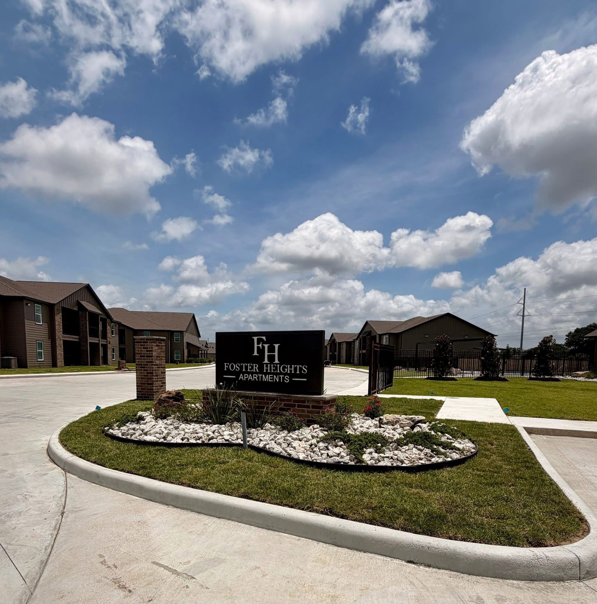 Entrance sign for a residential community with brick homes, landscaped flower bed, and a bright cloudy sky.