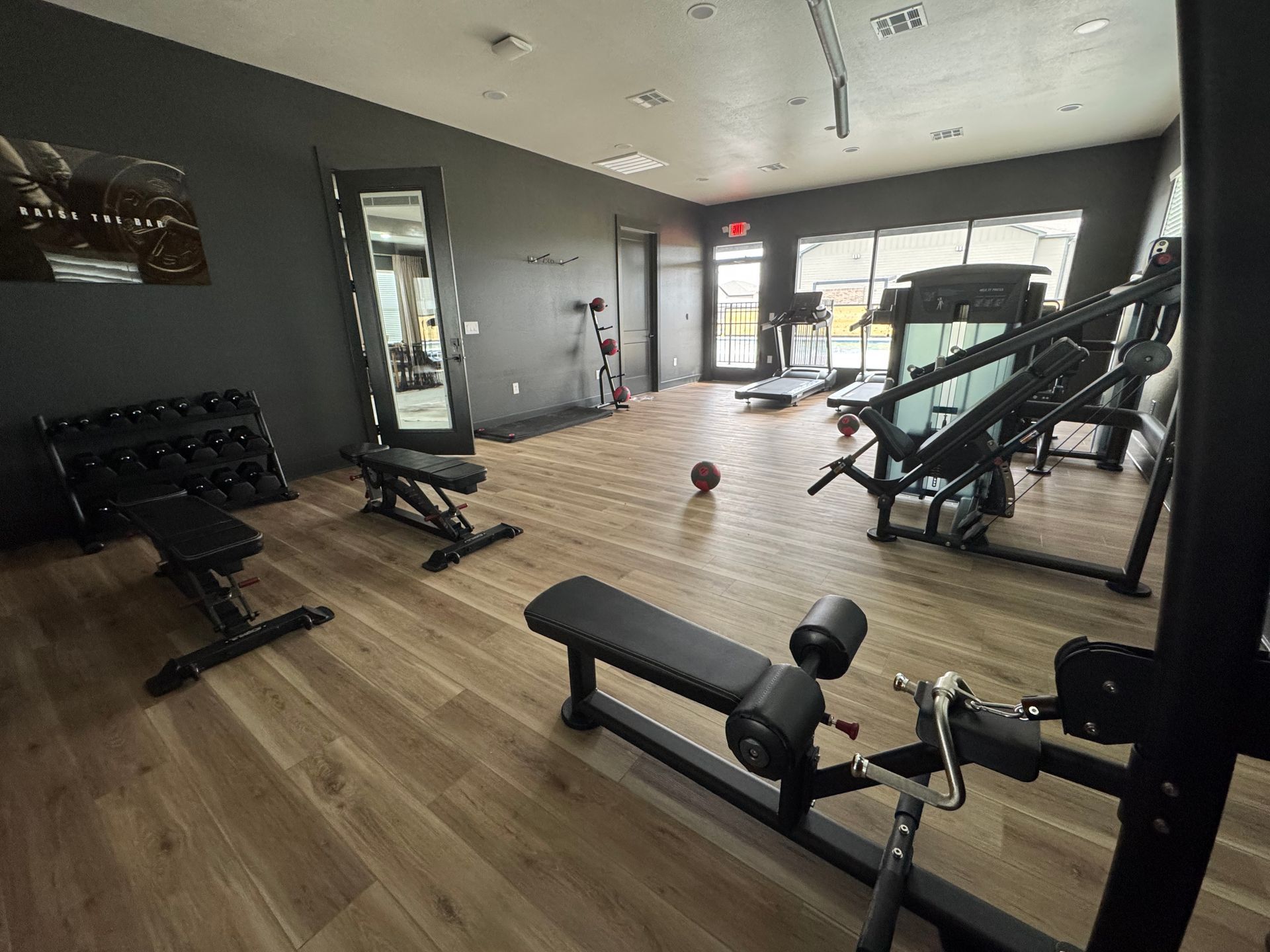 Empty gym with weight benches and exercise machines on a wood floor, lit by large front windows.