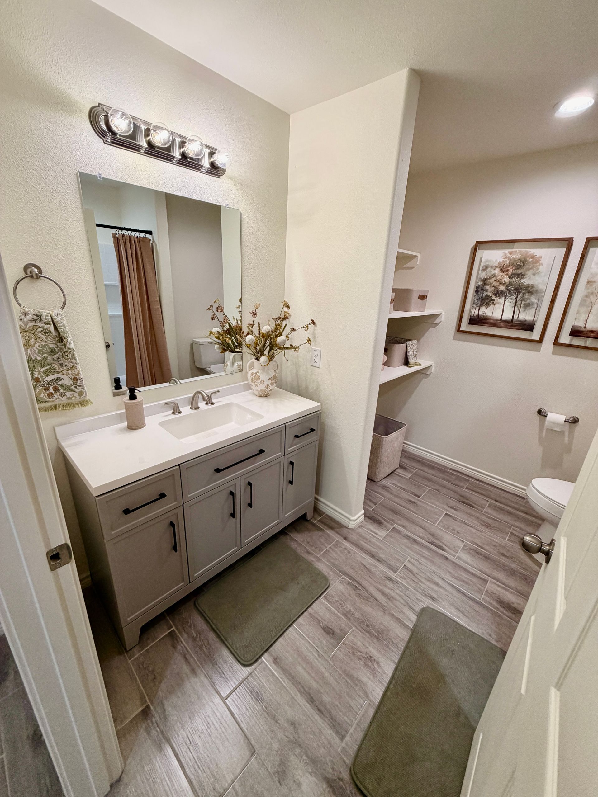 Bright bathroom vanity with mirror, sink, wall art, shelves, and green rugs on wood-look tile floor