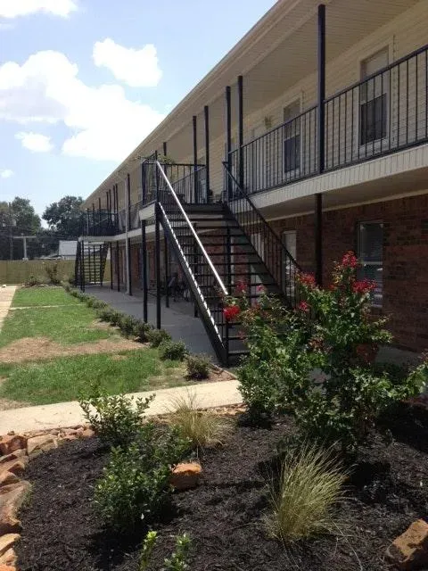 Exterior of an apartment building with black metal stairs, balconies, and landscaped shrubs under a blue sky