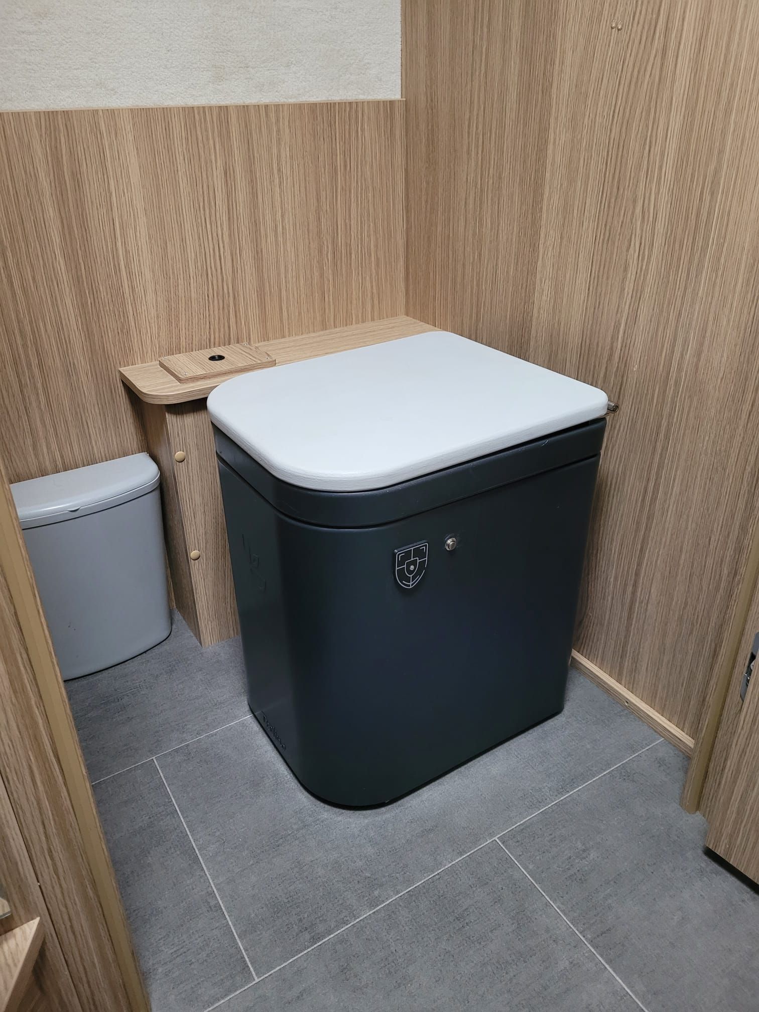 Dunkelgraue Toilette in einem kleinen Raum mit Holzvertäfelung und grauen Bodenfliesen.