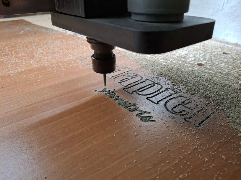 Eine CNC-Maschine schnitzt das Wort „Mpire“ in eine Holzoberfläche; Sägemehl ist sichtbar.