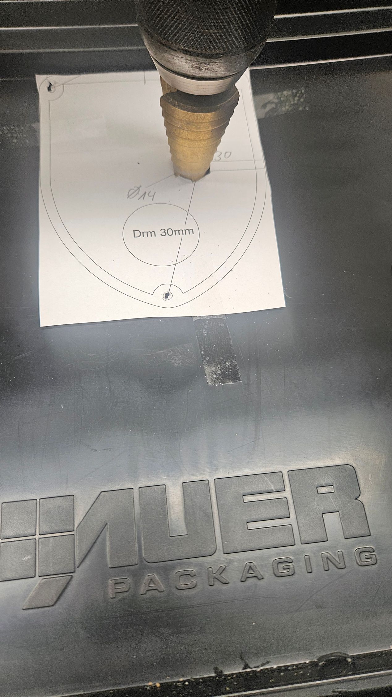 Eine CNC-Maschine schneidet eine Zeichnung in weißes Papier auf einer schwarzen Metalloberfläche; die Spindel der Maschine ist sichtbar.