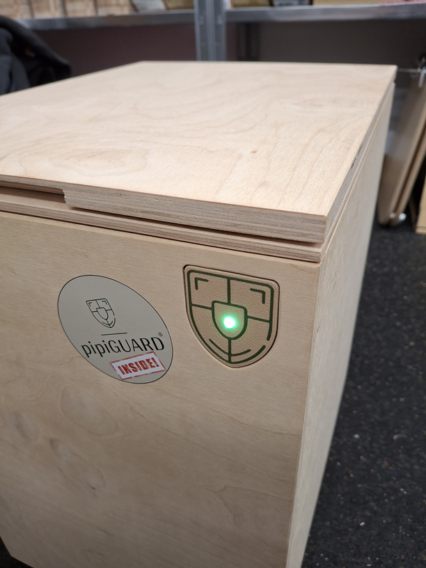 Holzkiste mit grünem Licht auf einem Schildlogo. Aufkleber „pipiGUARD“ an der Seite.