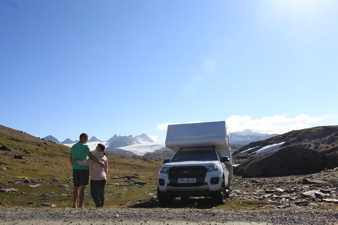 Ein Paar steht neben einem Wohnmobil und blickt auf schneebedeckte Berge unter blauem Himmel.