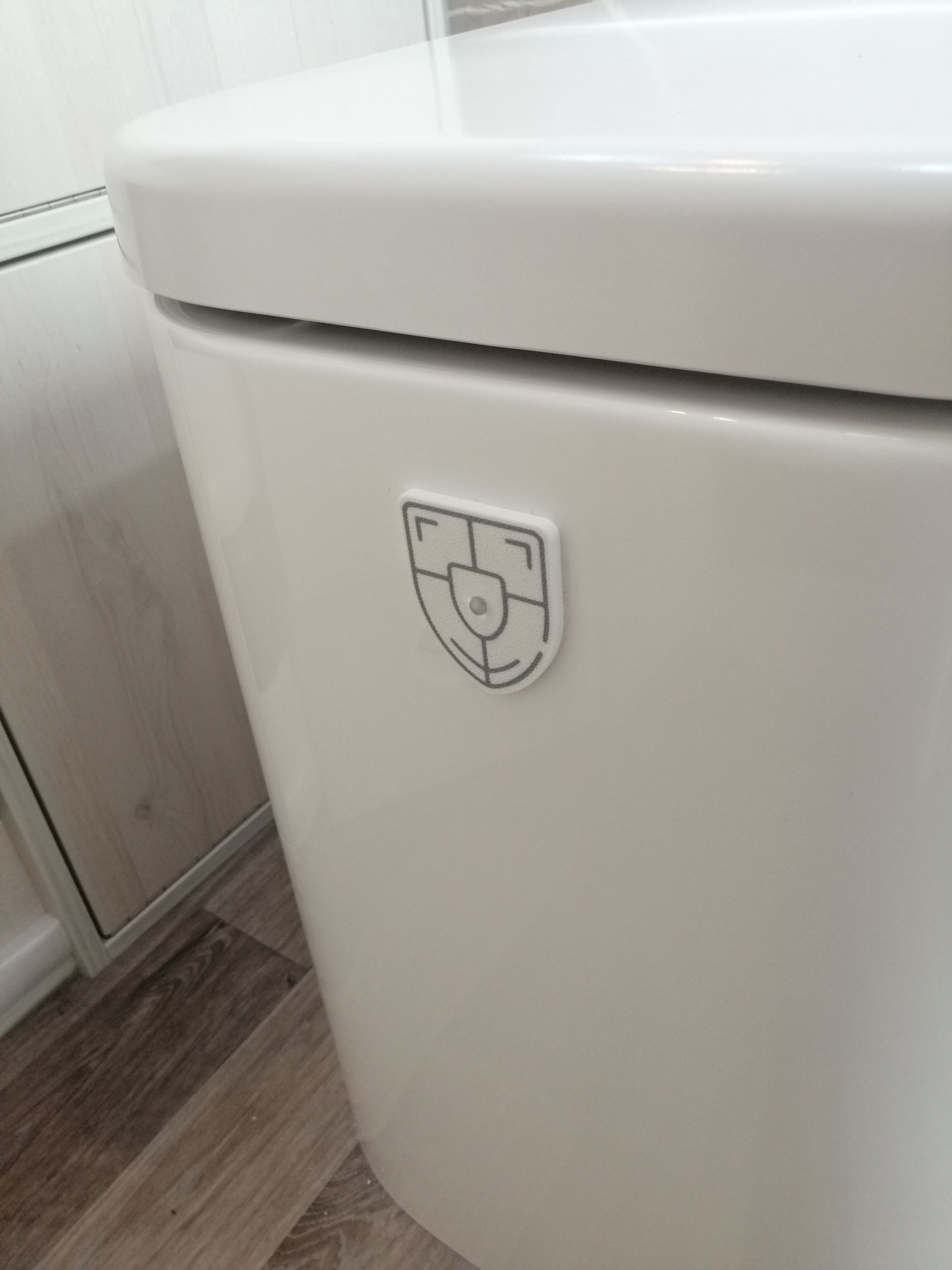 Weißer Spülkasten mit schildförmigem Logo an der Seite, in einem Badezimmer mit Holzbodenoptik.