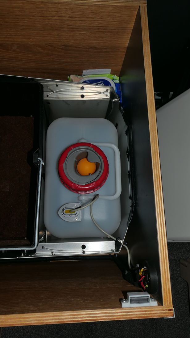 Ein grauer Kunststoffwassertank mit rotem Deckel in einem Holzschrank, mit Verkabelung und Digitalanzeige.