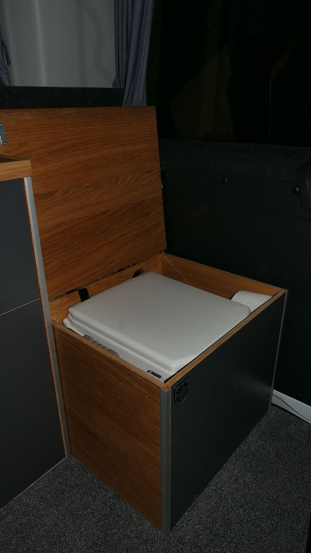 Holzkiste mit offenem Deckel, darunter befindet sich eine tragbare Toilette. Dunkelgraue Seiten, holzfarbener Deckel.