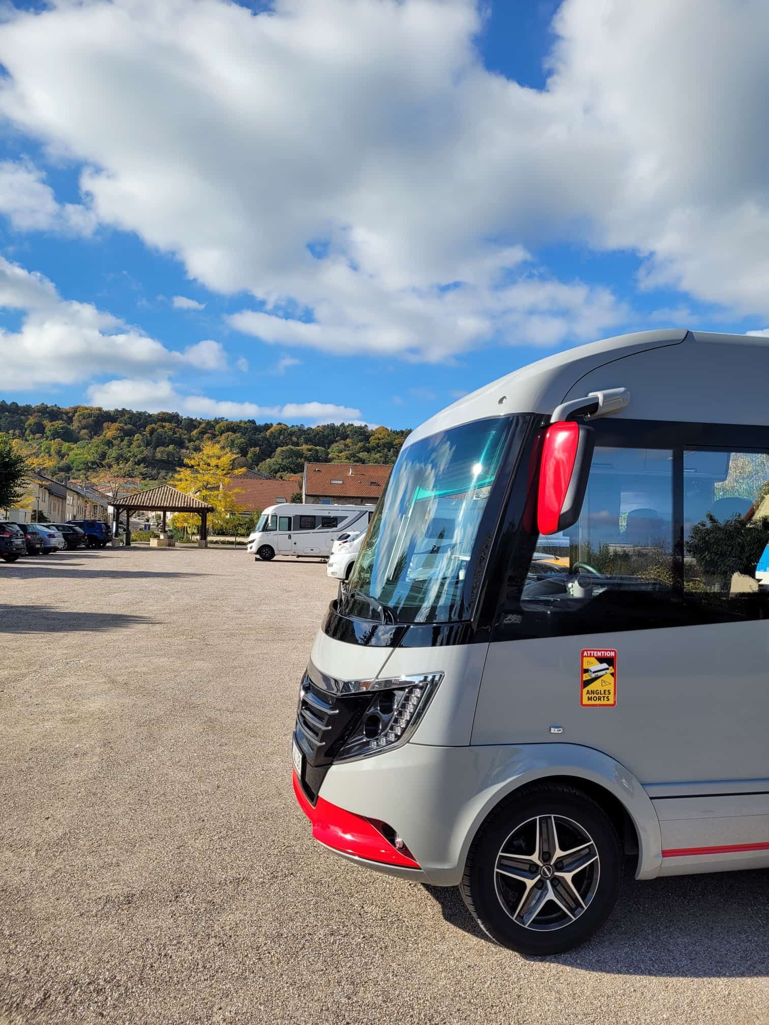 Ein grauer Campervan parkt an einem sonnigen Tag auf einem Schotterparkplatz. Rote Akzente am Fahrzeug.