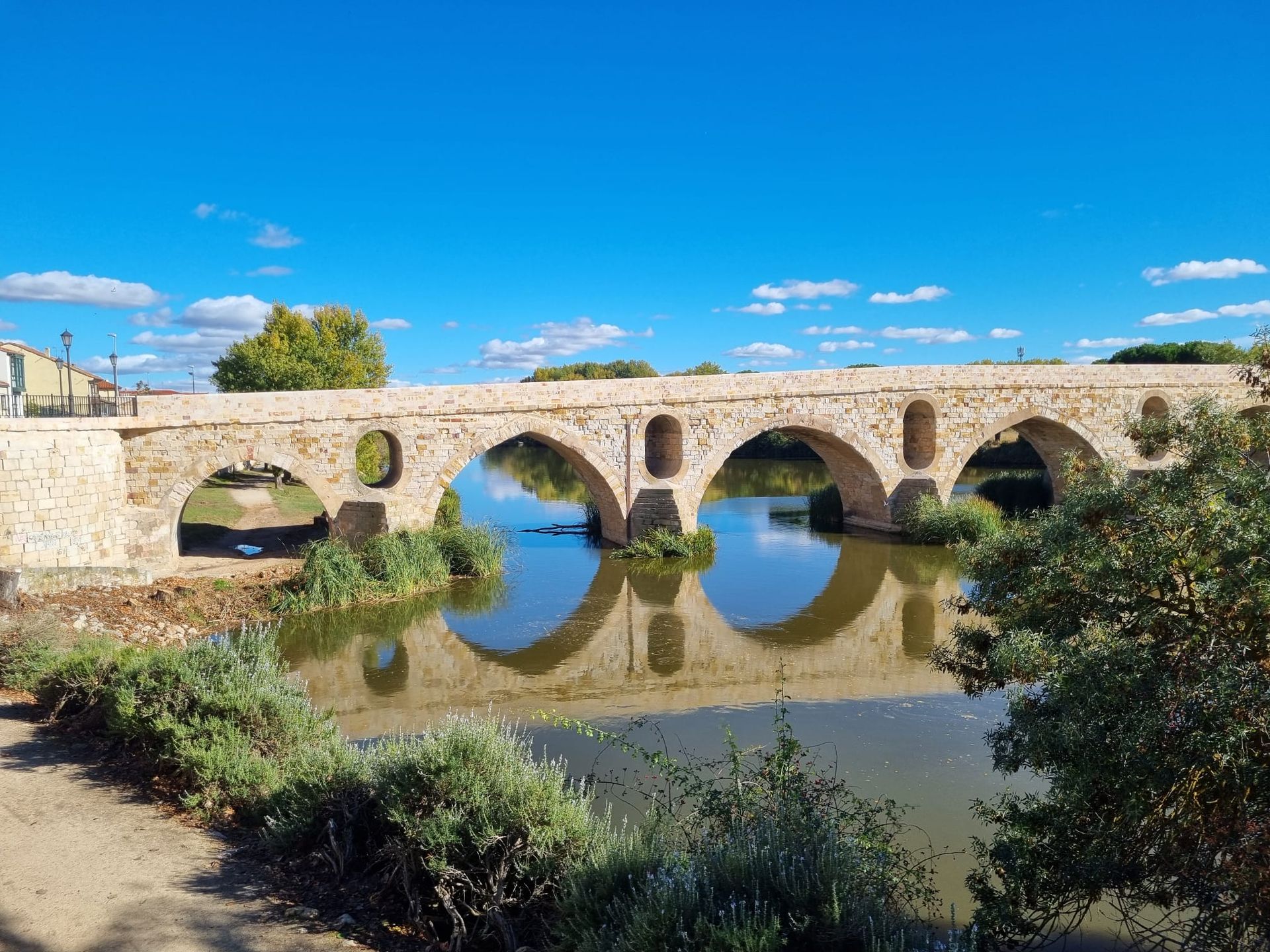 Steinbogenbrücke über ruhiges Wasser, die Bögen spiegeln sich unter blauem Himmel.