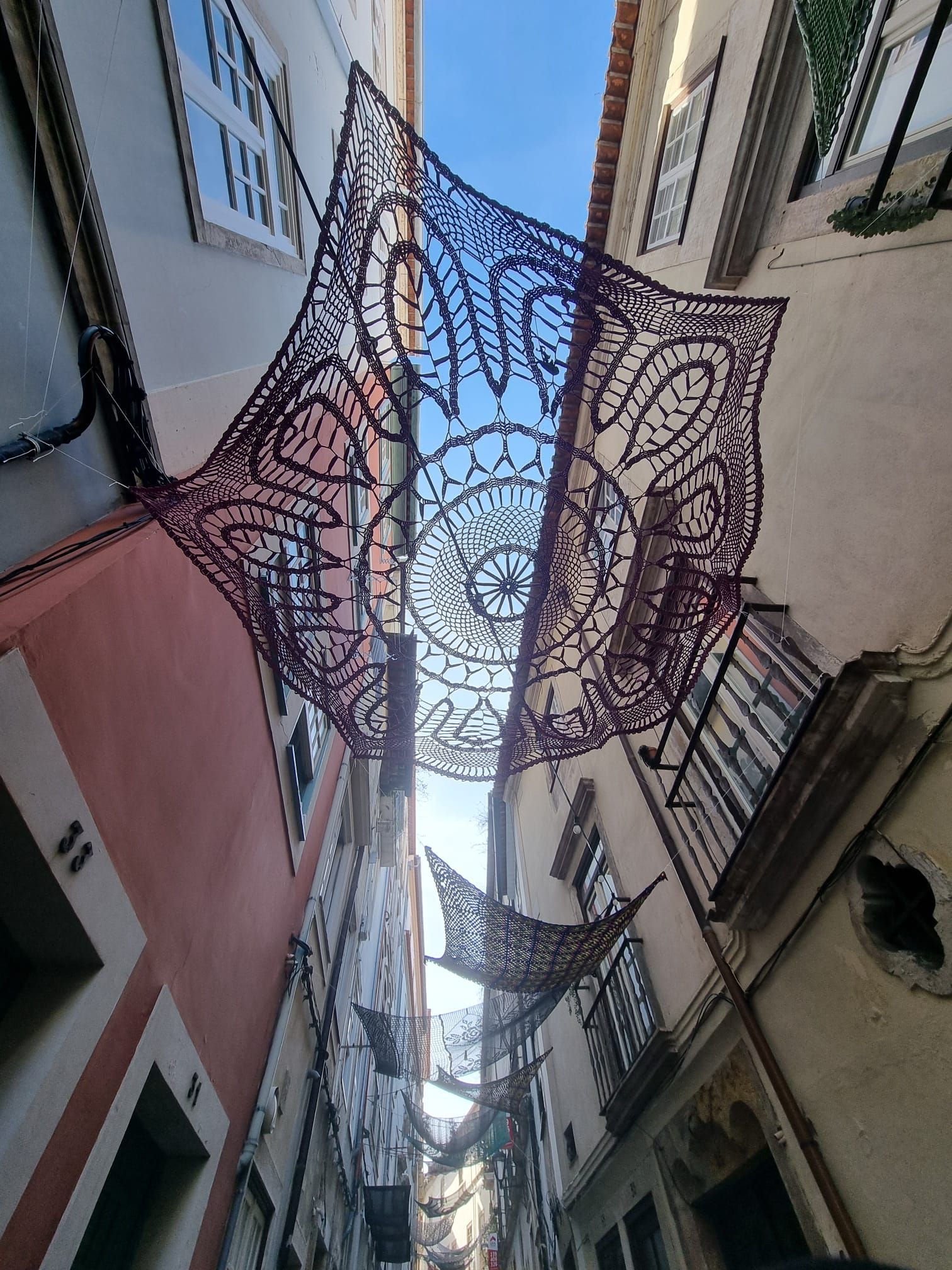 Eine Gasse mit dekorativer, lila Häkelkunst an der Decke, sonniger Himmel sichtbar.