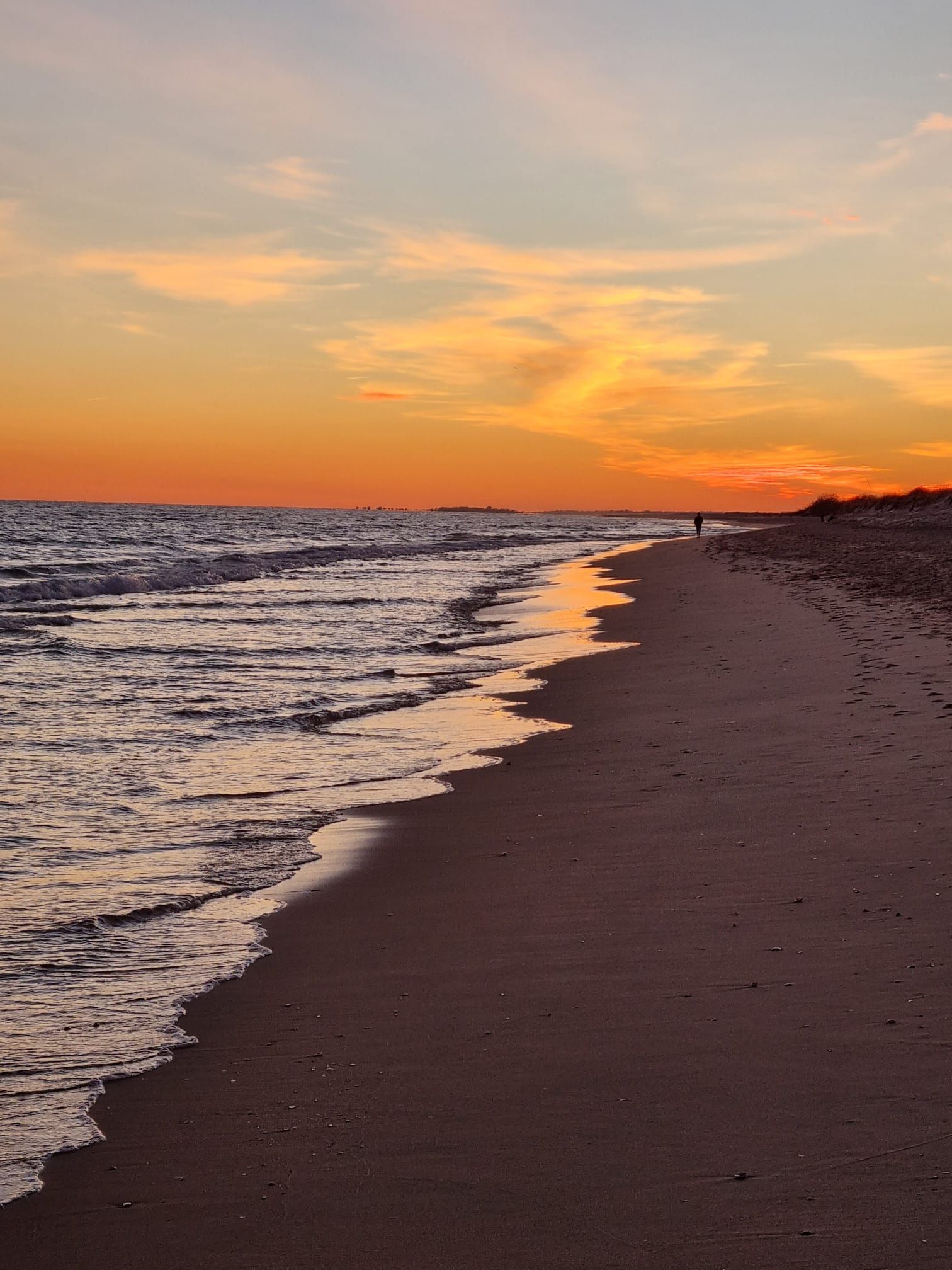 Strand bei Sonnenuntergang, orangefarbener Himmel, Wellen plätschern ans Ufer, eine Person geht am Strand entlang.