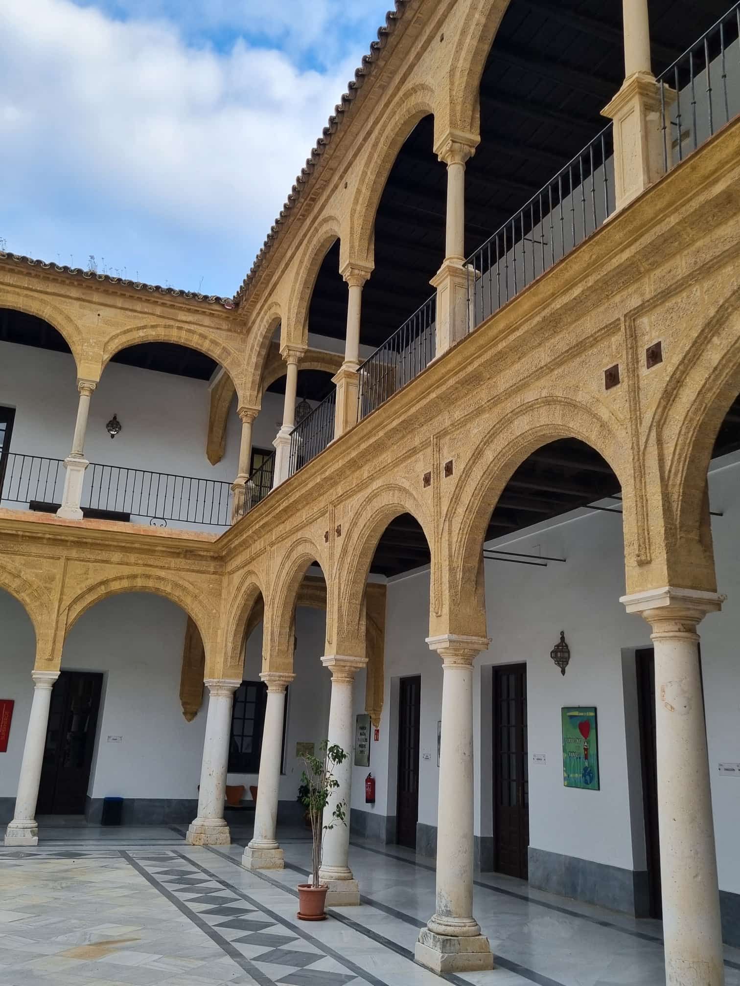 Innenhof mit Bogengängen, weißen Säulen und gemustertem Fliesenboden. Beige-weißes Gebäude unter bewölktem Himmel.
