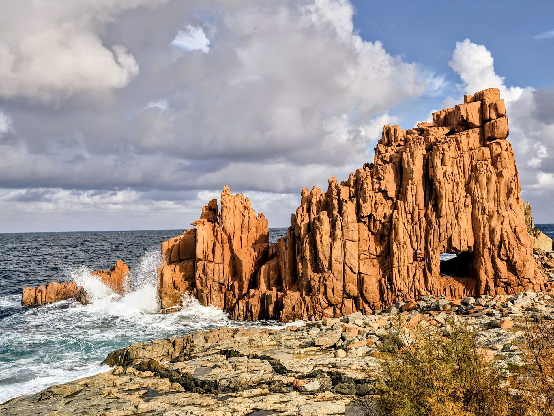Orangefarbene Felsformationen im Meer, an dem die Wellen brechen; bewölkter Himmel.