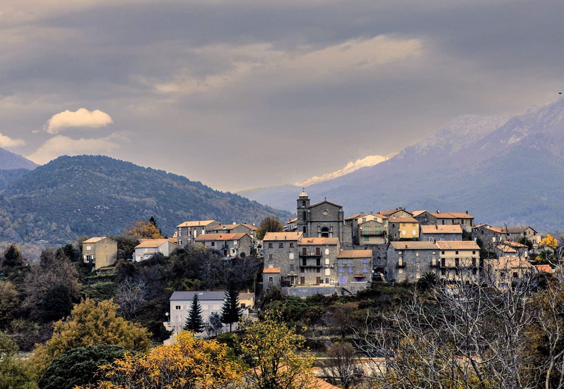 Ein Dorf auf einem Hügel mit Steingebäuden, einer Kirche und Bergen unter einem bewölkten Himmel.