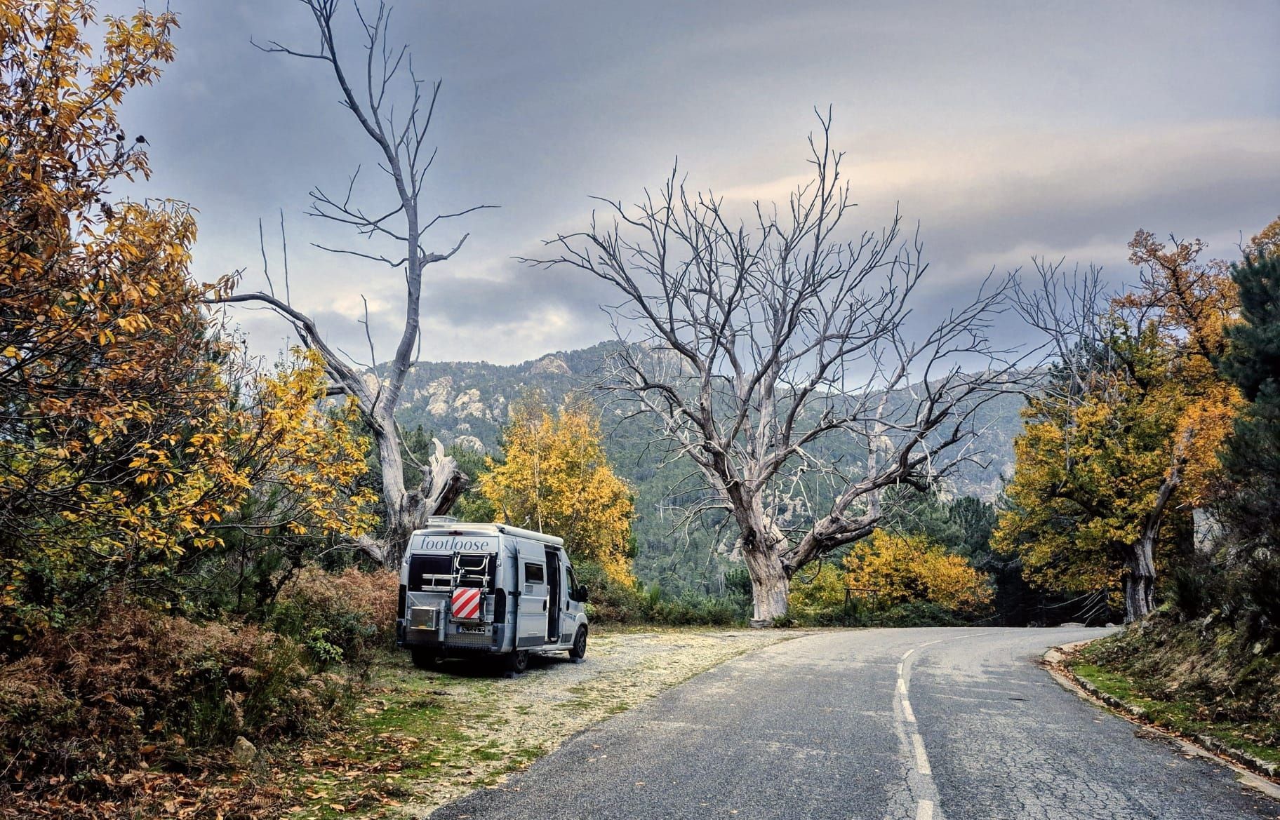 Ein Lieferwagen parkt im Herbst an einer malerischen Straße; Bäume mit gelben Blättern, bewölkter Himmel, Berge im Hintergrund.