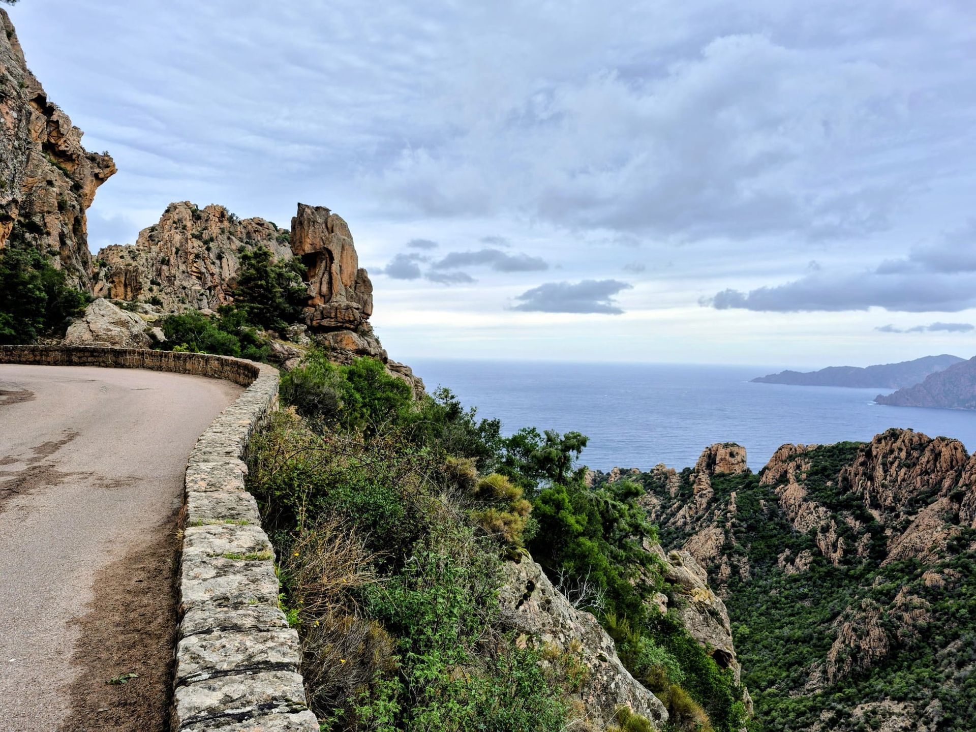 Kurvenreiche Küstenstraße mit Steinbarriere, Felsklippen, Meer und bewölktem Himmel.