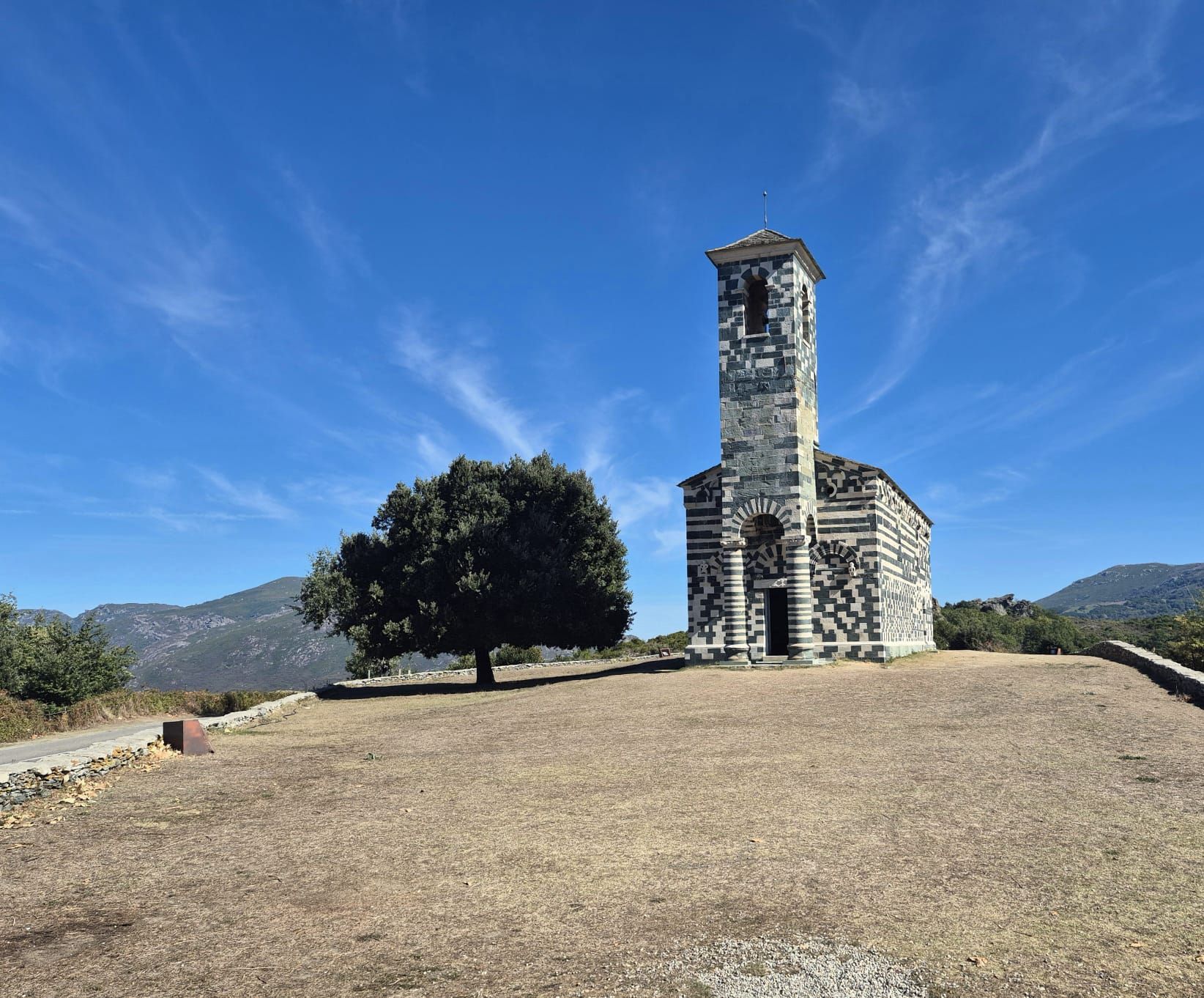 Steinkirche mit Glockenturm und Baum auf einem Hügel unter blauem Himmel mit Bergen in der Ferne.