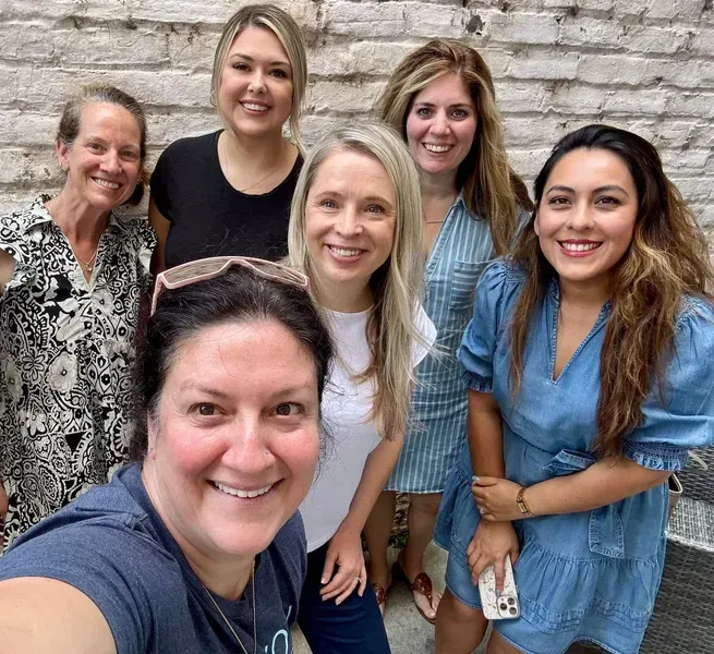 Six women smiling outdoors in front of a stone wall, posing closely for a selfie.