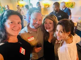 Four smiling people pose together at a warmly lit indoor social event.
