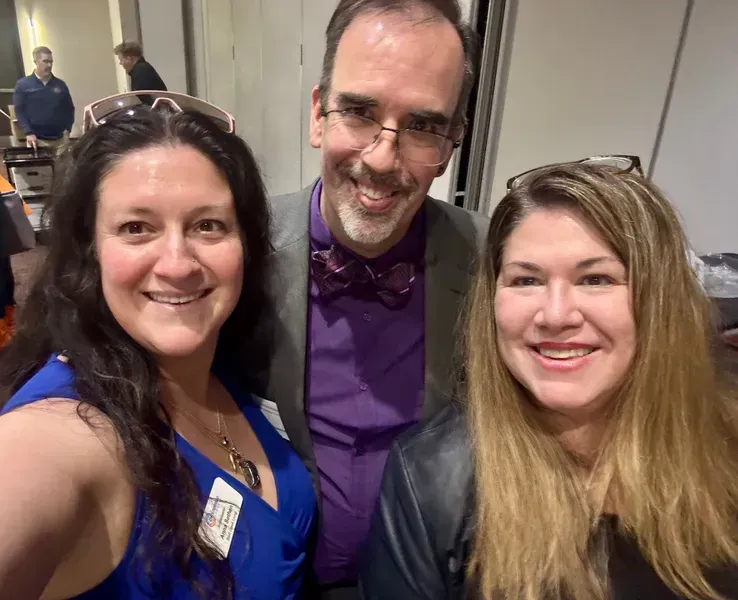 Three smiling people posing indoors at a conference, one in a blue dress, one in a purple shirt and bow tie.