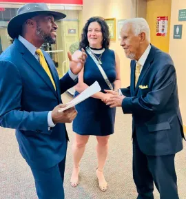 Three people in suits talking in a hallway, one holding papers and another smiling and gesturing.