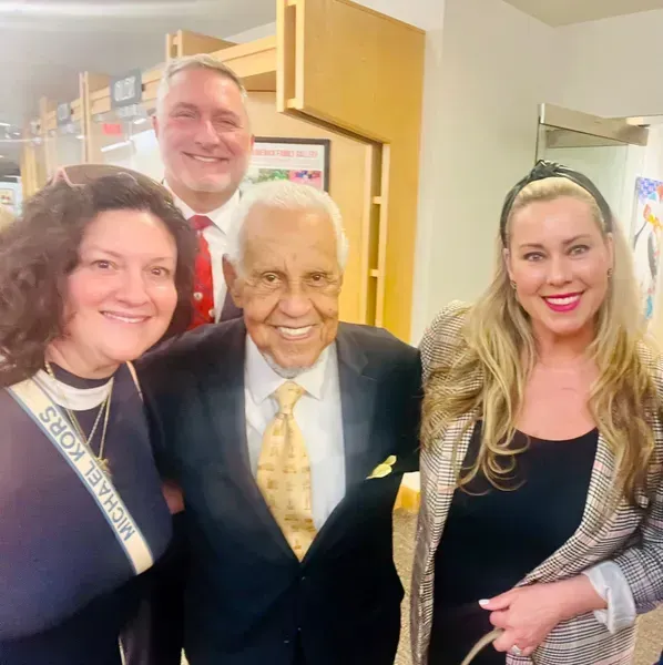 Four smiling people posing indoors, with an elderly man in a dark suit at center.