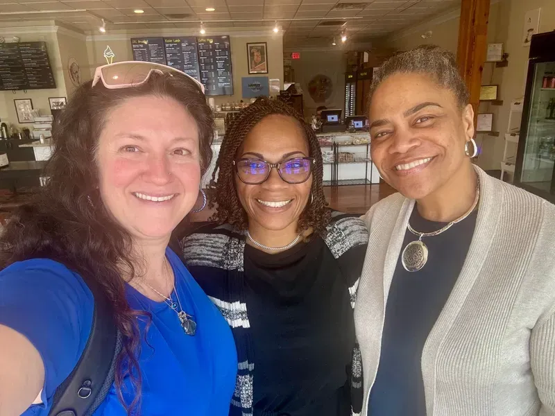 Three smiling people posing indoors, one holding the camera, in a cozy café.
