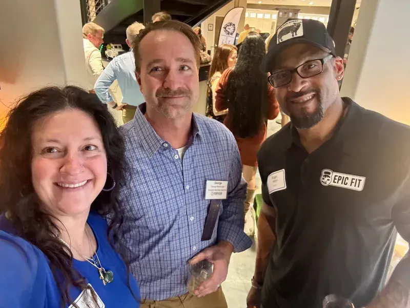 Three people smiling indoors at an event, with name badges and casual attire.