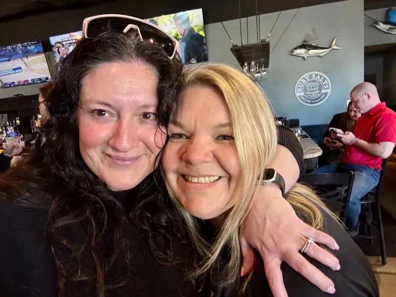 Two women smiling together in a restaurant, with wall decor and seated diners in the background.