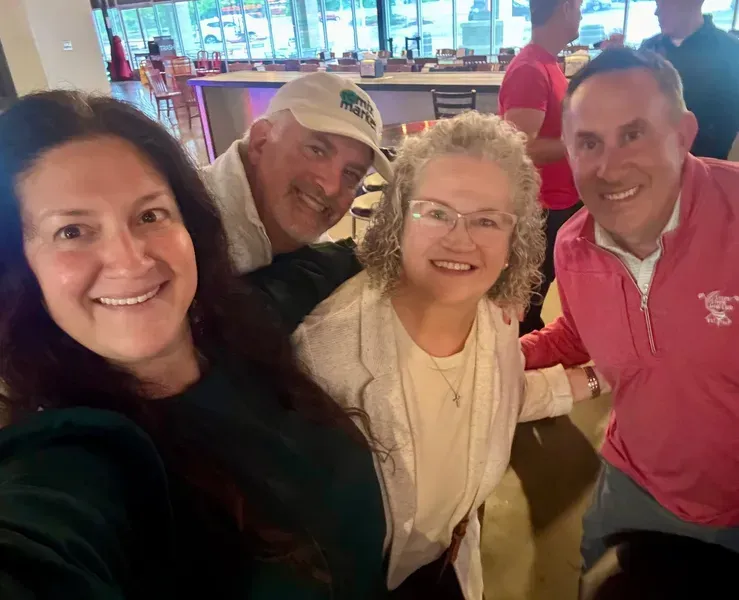 Four people smiling for a selfie in a bright indoor venue, with a bar and windowed background.
