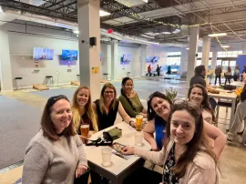 Group of friends smiling at a table in a bright indoor food hall, with drinks and plates in front of them.