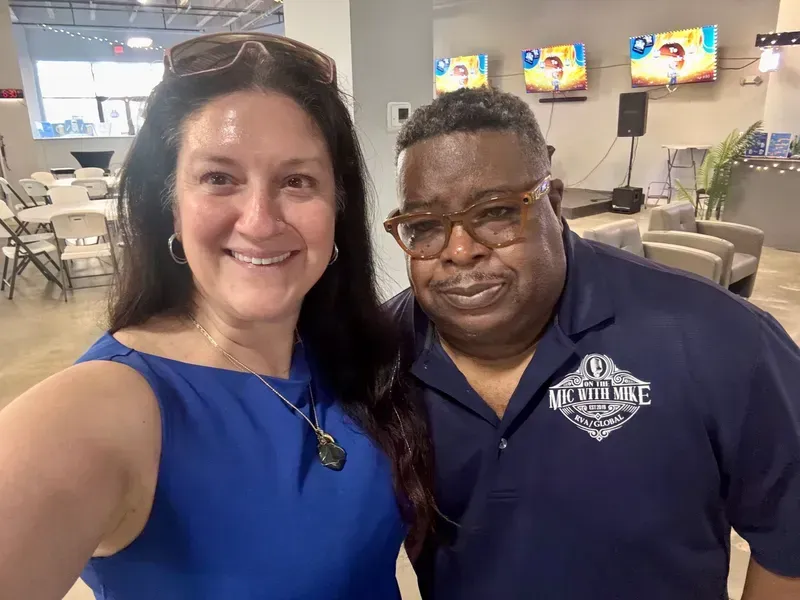 Two people smiling for a selfie in a casual dining hall with TV screens in the background.