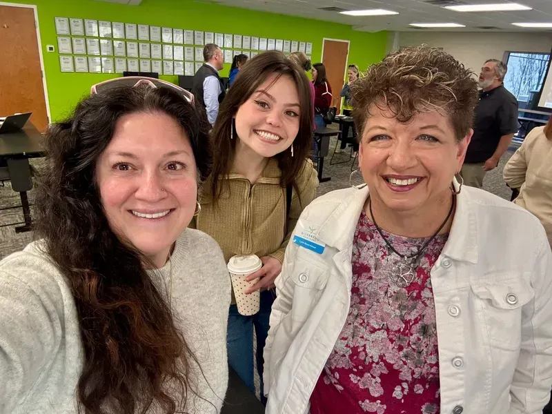 Three smiling people in a bright room with a green wall and papers displayed behind them.