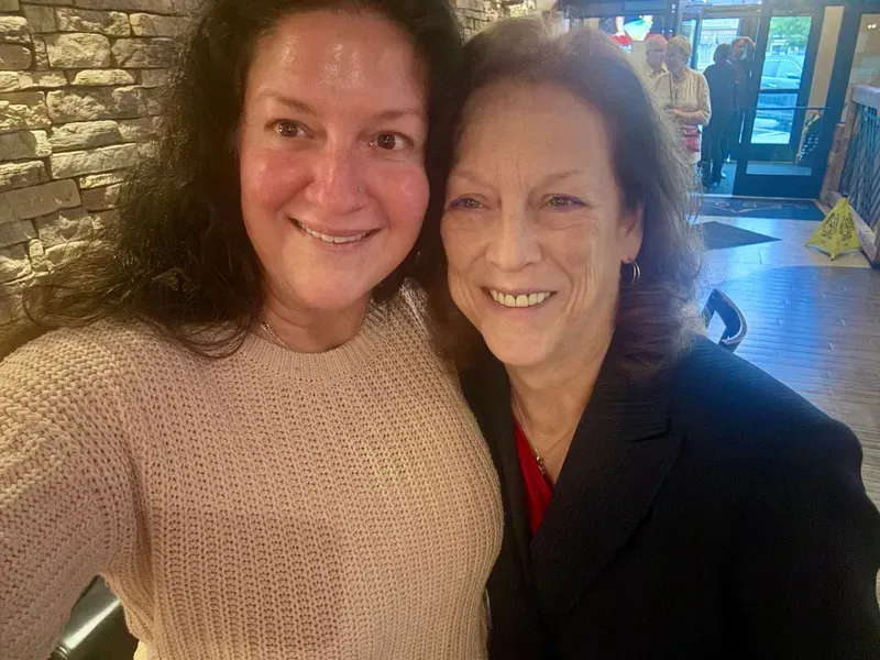 Two people smiling indoors, taking a selfie in a warmly lit lobby