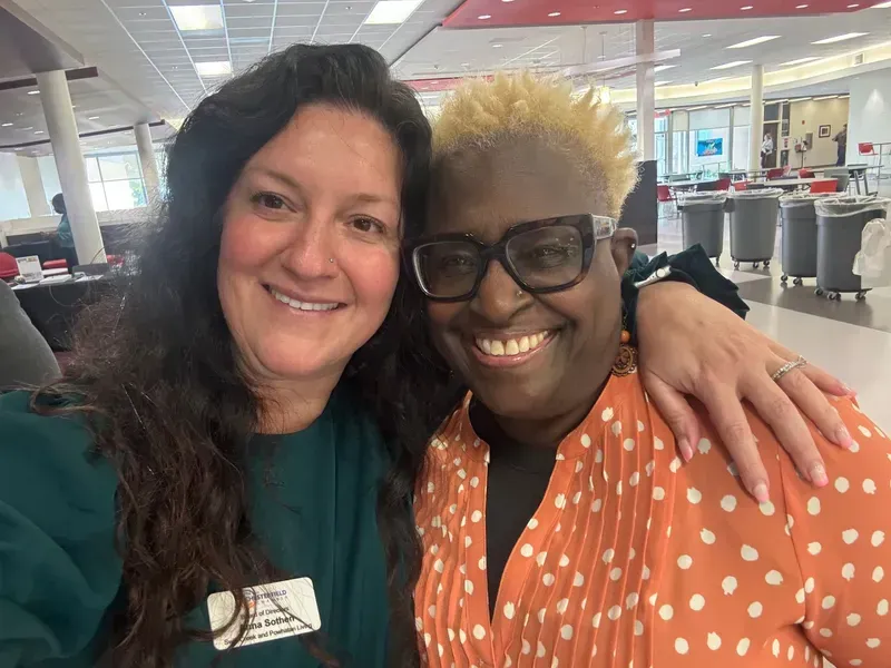 Two smiling coworkers taking a selfie in a bright office cafeteria