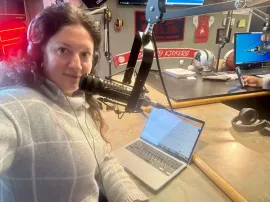 Person at a radio studio desk speaking into a microphone beside an open laptop.