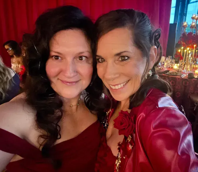 Two smiling people in red dresses posing together at a festive, candlelit event.