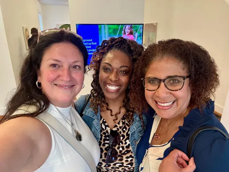 Three smiling people posing for a selfie indoors, with a TV visible in the background.