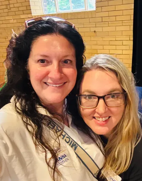 Two smiling people posing indoors, one in a white shirt and one wearing glasses, against a brick wall.