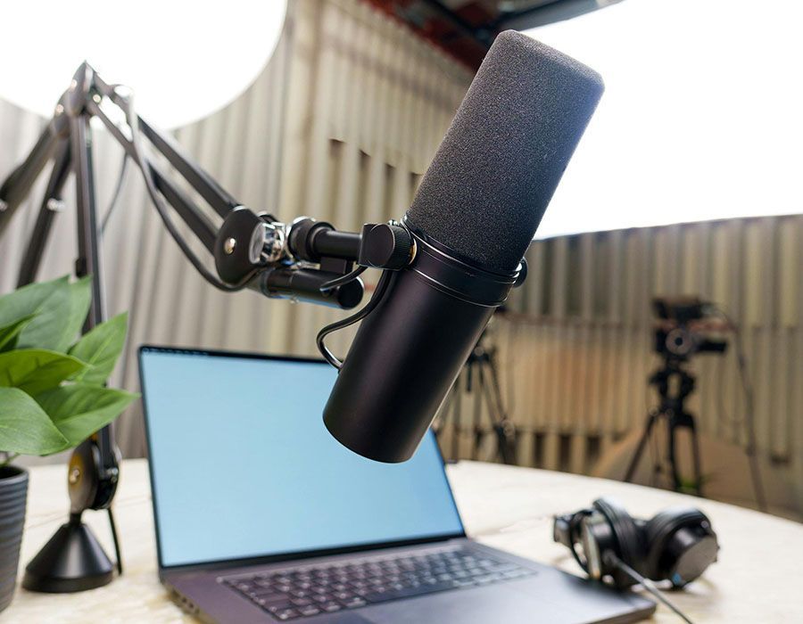 Podcast mic on boom arm over open laptop in a studio setup with headphones and a plant.