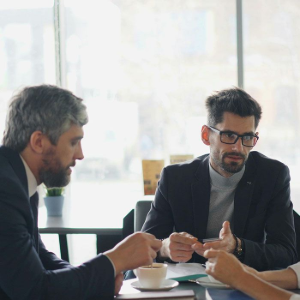 Two men in suits talk at a café table with coffee, one wearing glasses, in a bright office setting