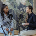 Two women in a podcast interview, seated with microphones and mugs against a floral mural backdrop