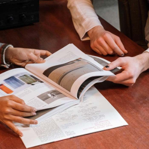 Hands reviewing an open magazine and paperwork on a wooden table