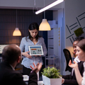Team meeting in a dim office, woman presenting a tablet while coworkers listen around a table.