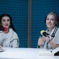 Two women speaking into microphones in a podcast studio with blue acoustic panels