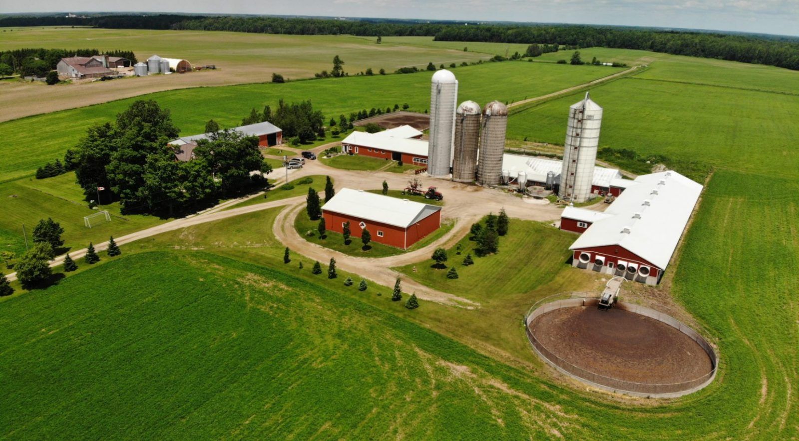 Farm scene with silo, barn, and green fields against a tree-covered hillside.