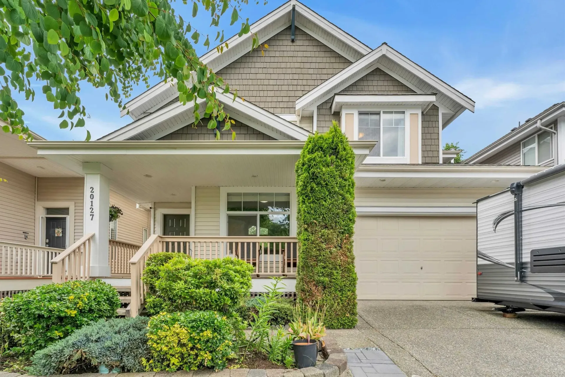 Suburban house exterior with front porch, beige siding, shrubs, and a garage driveway