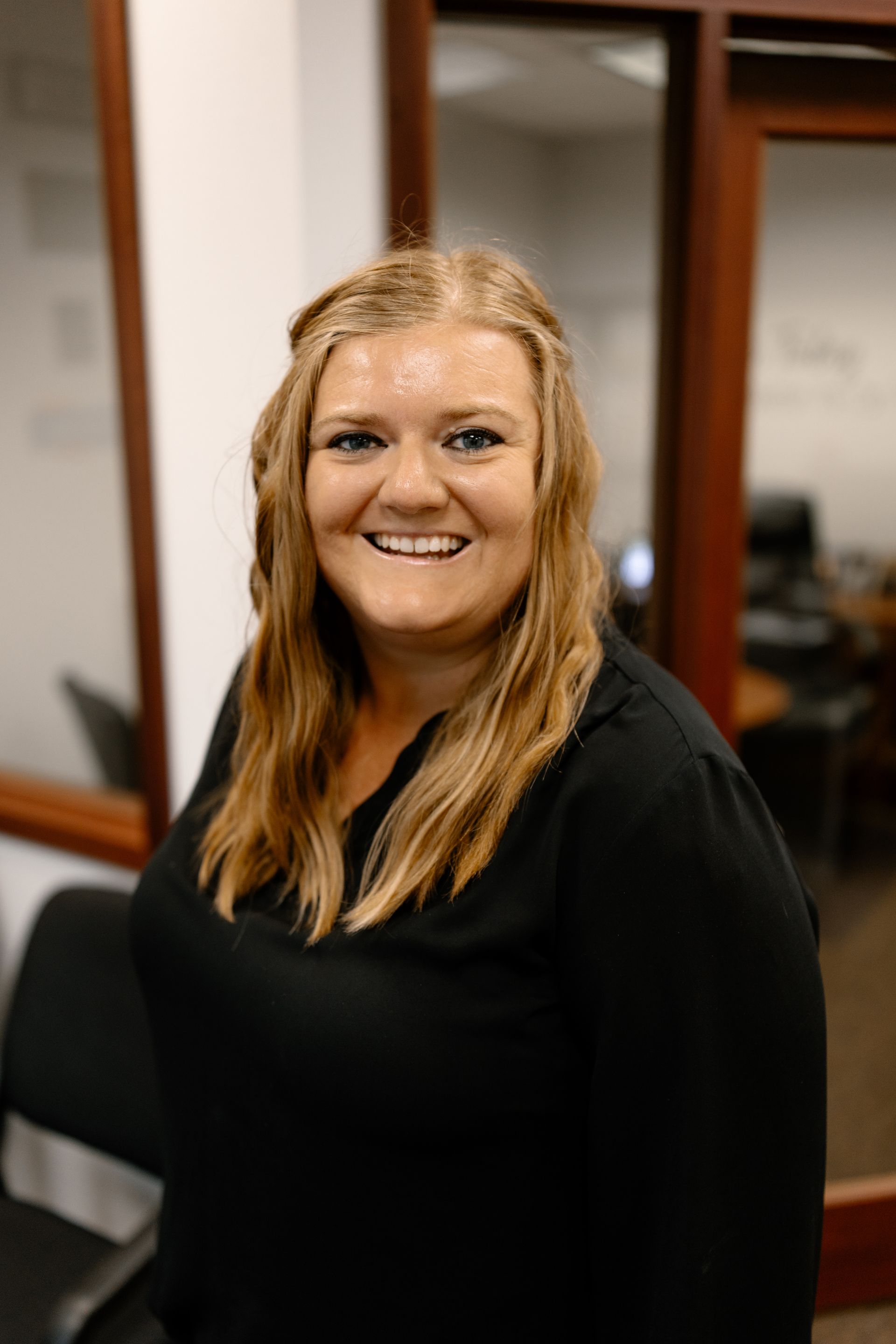 A smiling person with long light brown hair wears a black blouse in an office setting with glass partitions.