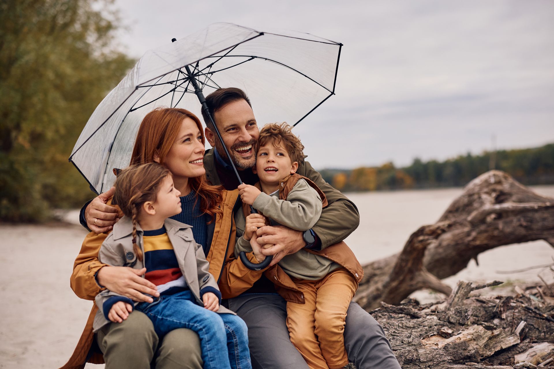 Family under umbrella by lake, embracing and smiling on overcast day.